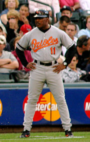 A man with dark skin stands on a baseball diamond. He is wearing a gray baseball uniform, which reads "Orioles" across the chest, and a black baseball helmet with a bird. He stands with arms akimbo and looks to the left of the image.