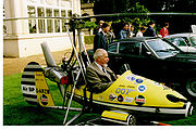 Small, one man, open-cockpit helicopter on a lawn about the size of a car next to it, with a man sitting in it.