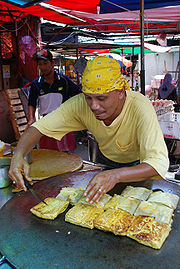 A cook making Murtabak, a type of pancake, in an outdoor stall. He is pictured leaning over his custom-made flattened wok filled with pieces of murtabak.