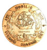 A round brass metal object features a sailing ship in the center over the U.S. Navy submarine service chest insignia of a dolphin and framed in a laurel wreath. In a circle around the edges are the Latin words "Ave Nobilis Dux, Iterum Factum Est" which translates to "Hail Noble Captain, It is Done Again."