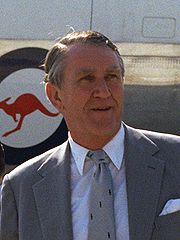 Colour photograph of a Malcolm Fraser aged about fifty, he has a weathered face and greying hair parted on the right. He wears a suit and tie; behind him can be seen part of a large aircraft with a kangaroo logo.