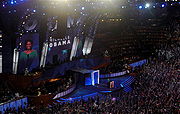 Michelle Obama speaks at a convention; her image and name are projected on a huge screen behind her. The large audience waves vertical blue signs.