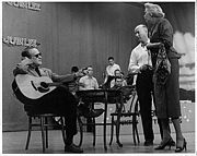 Red Foley sitting onstage at a table, left, with guitar on his lap; director Bryan Bisney and actress Fran Allison stand to the right
