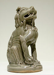 A small, green-grey statuette of a lion, sitting down and looking upwards. The lion's limbs are thin and angular.