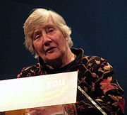 A middle-aged, white-haired woman with a dark pullover with flower pattern speaking from a podium.