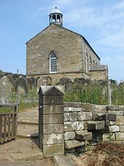 A small stone church standing in a churchyard on top of a hill.  On the near gable is a bell-cupola.