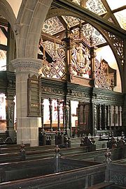 The interior of a church with an arch containing an elaborately carved screen in front of which are carved pews