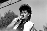 A black and white image of a light-skinned man with a microphone held to his mouth. He is visible from the chest up and wears a sleeveless black shirt with an opened sleeveless white vest overtop. A small cross is worn around his neck. His black hair is styled into a mullet. The man looks past the camera to the left. A mixture of trees and sky are visible in the background.