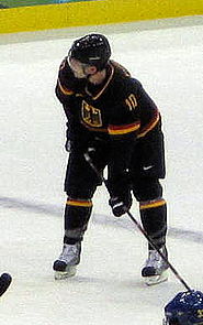 An ice hockey player dressed in a black and orange jersey watchfully skates in front of an opposing player who is playing the puck.