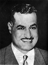 Head and shoulders of a man in his forties smiling. He has dark hair that is pulled back, a long forehead, thick eyebrows and a mustache.  He is wearing a gray jacket and a white shirt with a tie.