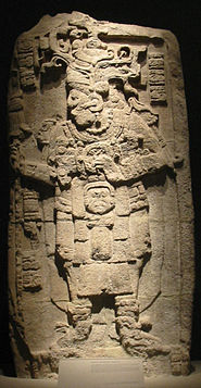 A relief sculpure showing a richly fressed human figure facing to the left with legs slightly spread. The arms are bent at the elbow with hands raised to chest height. Short vertical columns of hieroglyphs are postioned either side of the head, with another column at bottom left.