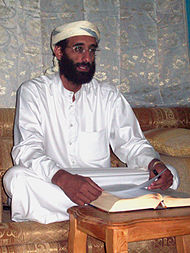 A man in white clothing with a beard and glasses sits cross-legged before a table with an open book.
