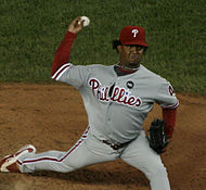 The same man as above in a gray baseball uniform and red baseball cap throws a baseball with his right hand from a pitcher's mound.