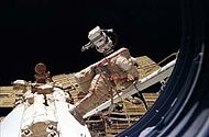 A man dressed in a white spacesuit with a red stripe manoeuvres along a boom-like crane towards a white cone-shaped space station module. Four arrays, one of which is damaged, project from the module, and the blackness of space forms the background. The rim of the porthole through which the photograph was taken is visible to the right of the image.