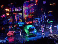 Bird's eye view of a stage, showing large scaffoldings, neon signs and a green car lying in the middle.