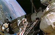 A man dressed in a white spacesuit with a red stripe seen clinging to the end of a boom-like crane, moving over a white space station module. Various trusses, solar arrays and other structures project from the module, and the Earth is visible in the background.