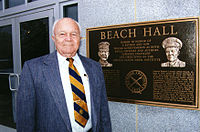Elderly man in coat and tie standing next to a bronze dedication plaque loacted at the building entrance.