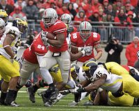 An American football player wearing scarlet and grey attempts to elude tacklers wearing Maize and Blue uniforms.