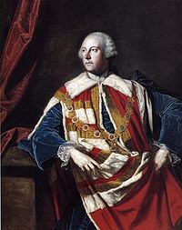 Man in his 50s wearing a white powder wig. He is wearing a decorative red and white robe over a blue jacket. He is looking to the left.