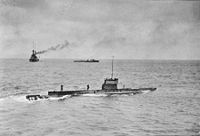 A submarine sailing on the surface of the ocean, while in the background is a large warship and a smaller vessel