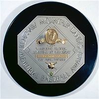 A black circle with an octagonal silver plaque in the middle. The edge of the plaque reads "KENESAW MOUNTAIN LANDIS AWARD BASEBALL MEMORIAL". In the middle of the octagon is a baseball diamond which contains, from the top, a man's face in gold, "Most Valuable Player", the winner's league, his name in a gold rectangle, and his team.