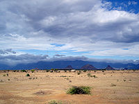 Daytime view of an almost lifeless expanse, dry rocks and sand marked only by the odd lone shrub. The dry terrain reaches to a chain of mountains in the far distance, near the horizon. A bank of clouds soars above the void, but it does not appear to hold the promise of rain. A far darker, larger, more turgid cloud bank sits above the distant mountains, above the horizon.