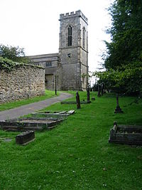The east end of a a stone church is seen through a graveyard from the north.  On the right is a battlemented tower and the nave extends to the left