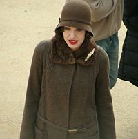 From waist–up, a pale skinned, red-lipped slender woman in a cloche hat and fastened fur-trimmed coat, both brown