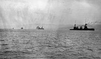 Four warships at sea sailing in line ahead, the shoreline distant in the background.