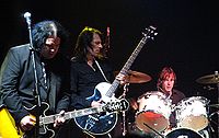 Prominent in the foreground, two guitarists concentrate on their playing, while the drummer, a little behind them to their left, toils away.