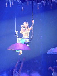 A female brunette performer. She is singing while suspended on a giant umbrella in the air. She wears eastern-inspired clothes.