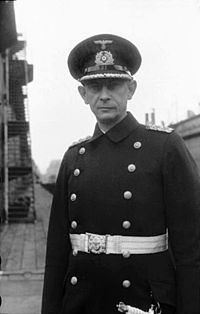 The head and upper torso of a man. He wears a peaked cap, black naval coat and a white belt with dagger. His facial expression is determined; his eyes are looking straight into the camera.