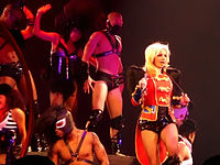 Image of a blond woman. She is standing with a red feathered jacket, carrying a whip around her neck and singing in a wireless microphone. Several people surround her, all wearing S&M outfits.