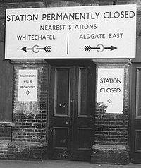 A narrow wooden door between two brick buttresses. A large sign above saying "Station Permanently Closed, Nearest Stations Whitechapel/Aldgate East". Arrows point left and right to each of the two stations named.