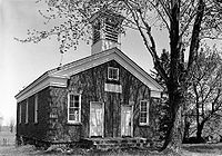 A black and white image of a building surfaced in small round stones, seen from slightly to its left. It has a pointed roof with a square cupola on top in the front. A large tree is in front of it, partially obscuring the view