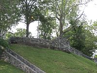 Photograph of a crumbling brick wall running up and then alongside a grassy hill which has trees and a lamppost at the top. Brick buildings are visible in the background to the left.