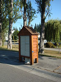 A community letterbox with a cedar shelter built over it. The box is located on a residential sidewalk in front of a small park.