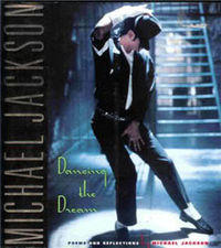 Front cover of book showing a man under a spotlight in front of metal stairs. He wears black trousers, a black shirt and a black fedora. Under his shirt is a white T shirt, which matches the color of his socks, right armband and right arm brace. The man is striking a pose: legs apart and to the left, he looks down to the ground, as his braced right hand holds his hat atop his head. His left hand covers his crotch.