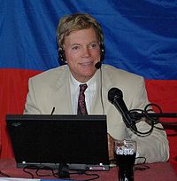 A photographic portrait of a blond-haired, green-eyed man who is wearing a suit and tie and is about 50 years old. He is wearing a headset, speaking into a microphone, and sitting in front of a table. On the table is a Coca-Cola glass and a laptop computer.