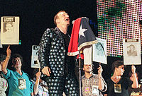 A light-skinned man with brown hair singing into a microphone on a stand, which has a flag draped over it. His shirt and trousers are both grey and feature a design of many overlapping circles. He faces to the right. A line of women stand behind him, each one holding up a sign that says "Donde Estan" or "Judicia". Every sign has an image of a different person below the text.