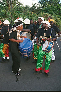 Band members wearing black t-shirts and white hats play drums in the street. Tropical foliage in the background.