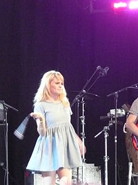 Image of a young, blonde woman performing on stage. The woman is wearing a short, light blue dress. The performer is seen to be swinging a microphone.