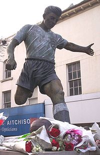 A statue of a man wearing a white shirt and blue shorts, about to kick a football