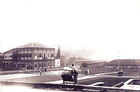 A photograph showing workers spreading or gathering coffee beans drying on a large paved plaza with an elegant, two story neoclassical building on the left and warehouses and other plantation buildings in the background