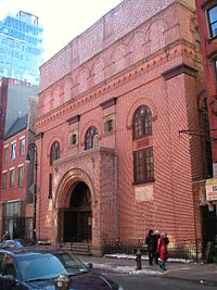 A five-story square building directly abuts a sidewalk. The facade is reddish brick, with two square windows on the second and three arched windows on the third floor. The main entrance juts forward from the facade, and is topped by an arch.