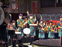 A group of eight young people wearing plaid kilts, green shirts, knee-high socks and navy berets stand on stage in a line, four playing bagpipes, four playing drums