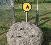 Boulder carved with the words "Für die Opfer der Unmenschlichkeit". In the background are a section of border fence and a yellow sign showing a kneeling soldier taking aim with a rifle.