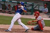 A man in a baseball uniform with white pants and a blue shirt swings while batting. The catcher in red and gray is also pictured.