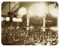 A photograph showing a large number of men seated on semi-circular tiers in a vaulted chamber as a large crowd looks on from an arcaded balcony