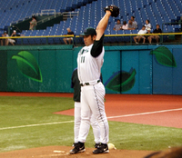 A man stretches his arms behind his head while wearing a baseball glove and a white baseball uniform with green sleeves and cap.